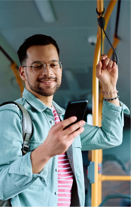 Homem jovem de barba e óculos, sorrindo enquanto utiliza o aplicativo da Exmed no seu smartphone e se apoia em uma alça de segurança dentro de um ônibus.