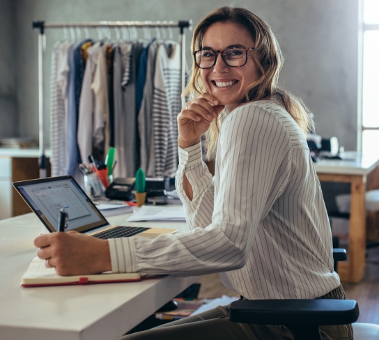 Mulher sorridente de óculos escrevendo em um caderno, com um laptop com o Exmed Empresas à frente e roupas penduradas ao fundo.
