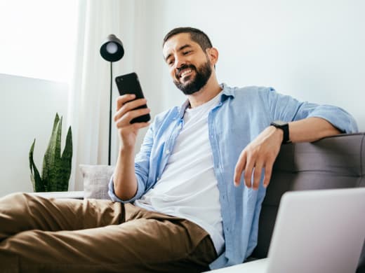 Homem sorridente sentado no sofá, usando o smartphone com plantas ao fundo.